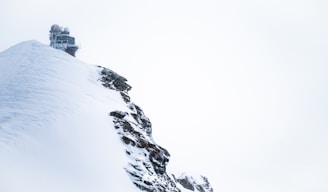 A scientific research station or observatory perched on the snowy peak of a mountain. The structure is surrounded by snow and ice, with rocky elements visible on the edge of the ridge. The sky is overcast and mostly white, blending with the snowy landscape.