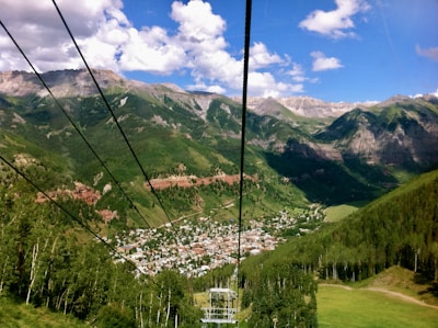 A scenic view of a small town nestled in a lush green valley surrounded by majestic mountain ranges. The town has white and red buildings and is accessible via elevated cables, suggesting a ski lift or gondola. The mountains in the background are partially covered with trees, and the sky is bright with scattered white clouds.