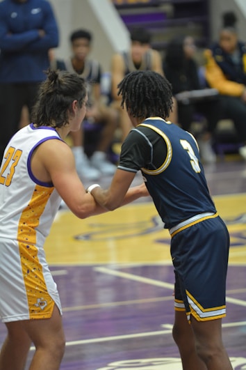 Two basketball players are interacting on a court, with a focus on their jerseys numbered 23 and 3. The players are in motion, engaged in a dynamic play. In the background, other team members sit on the benches dressed in matching uniforms, with blurred figures suggesting spectators or coaching staff.