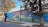 A mural painted on the side of a building featuring a whimsical scene with vibrant imagery. There are large trees integrated into the design, with a person reading and another climbing a branch. The artwork includes rich detail with elements like flowers and a picturesque horizon.