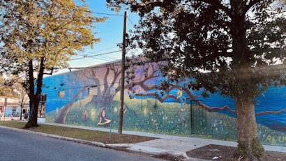 A mural painted on the side of a building featuring a whimsical scene with vibrant imagery. There are large trees integrated into the design, with a person reading and another climbing a branch. The artwork includes rich detail with elements like flowers and a picturesque horizon.