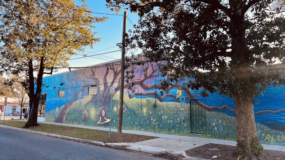 A mural painted on the side of a building featuring a whimsical scene with vibrant imagery. There are large trees integrated into the design, with a person reading and another climbing a branch. The artwork includes rich detail with elements like flowers and a picturesque horizon.