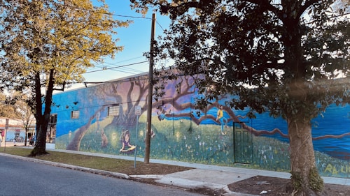 A mural painted on the side of a building featuring a whimsical scene with vibrant imagery. There are large trees integrated into the design, with a person reading and another climbing a branch. The artwork includes rich detail with elements like flowers and a picturesque horizon.