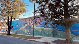 A mural painted on the side of a building featuring a whimsical scene with vibrant imagery. There are large trees integrated into the design, with a person reading and another climbing a branch. The artwork includes rich detail with elements like flowers and a picturesque horizon.