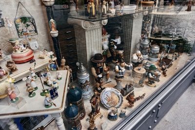 The image displays a shop window filled with various miniature figurines, ornaments, and decorative steins. There are wooden figures dressed in traditional Bavarian attire, along with colorful children figurines holding umbrellas and flowers. Some ceramic and metal steins decorated with German motifs are visible. The window reflects a cobblestone street outside.
