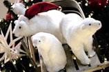 Three plush polar bear toys are set against a festive background with Christmas decorations. One of the bears is wearing a red Santa hat, and they are positioned on a sled. Greenery and lights add to the holiday atmosphere.