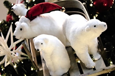Three plush polar bear toys are set against a festive background with Christmas decorations. One of the bears is wearing a red Santa hat, and they are positioned on a sled. Greenery and lights add to the holiday atmosphere.