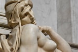 A marble sculpture of a seated figure with detailed facial features and a contemplative expression. The sculpture has draped fabric around the head and shoulders, showcasing intricate carving. The marble has a smooth texture with subtle variations in color, indicative of classical art styles.