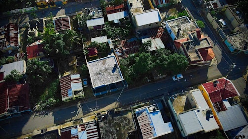 An aerial view of a residential neighborhood with various houses featuring flat and sloped roofs. The area is lush with greenery, and the houses are arranged closely together, divided by a narrow road where a few vehicles are parked. The rooftops vary in color and material, including red tiles and gray concrete.