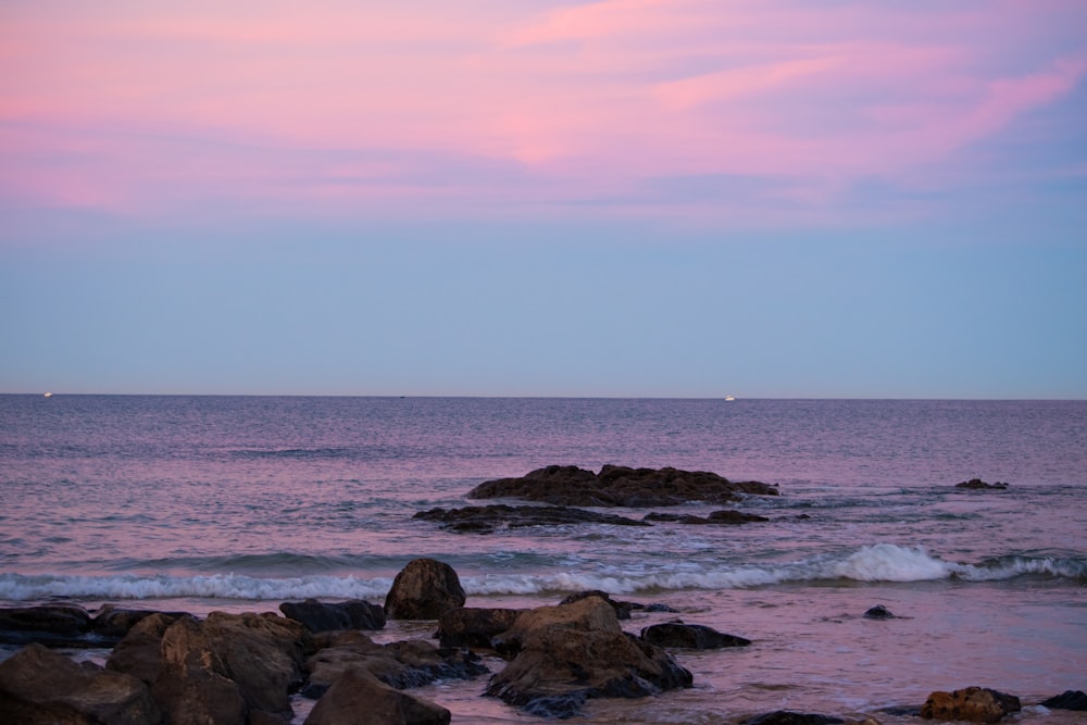 a pink sky over the ocean with rocks in the foreground