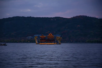 A large, ornate boat with traditional architectural features sails on a calm body of water. The boat is adorned with gold details and resembles a traditional Chinese dragon boat, complete with dragon head and tail on either end. In the background, a hilly landscape cloaked in dusk light creates a serene setting.