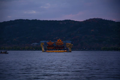 A large, ornate boat with traditional architectural features sails on a calm body of water. The boat is adorned with gold details and resembles a traditional Chinese dragon boat, complete with dragon head and tail on either end. In the background, a hilly landscape cloaked in dusk light creates a serene setting.