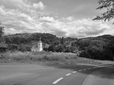 A serene countryside scene features a small white church with a steeple nestled among lush green hills. A winding road leads through the landscape, bordered by wild grasses and flowers. The sky is partly cloudy, adding depth to the picturesque view.