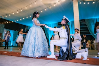 A young woman in an elaborate light blue gown stands on a red carpet inside a decorated venue. She is being approached by a man dressed in a formal military-style uniform, kneeling in front of her with a cape. Several other people are present in the background, observing the scene. The venue is adorned with string lights and blue drapery, contributing to a festive atmosphere.