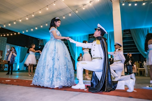 A young woman in an elaborate light blue gown stands on a red carpet inside a decorated venue. She is being approached by a man dressed in a formal military-style uniform, kneeling in front of her with a cape. Several other people are present in the background, observing the scene. The venue is adorned with string lights and blue drapery, contributing to a festive atmosphere.