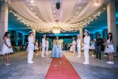 A formal event featuring a girl in an elegant blue gown standing on a red carpet, surrounded by uniformed individuals in white suits with hats. The setting is beautifully decorated with draped white fabric on the ceiling and an abundance of string lights contributing to a festive atmosphere. A chandelier hangs centrally, illuminating the scene. Several other people dressed in formal attire stand on either side, observing the moment.