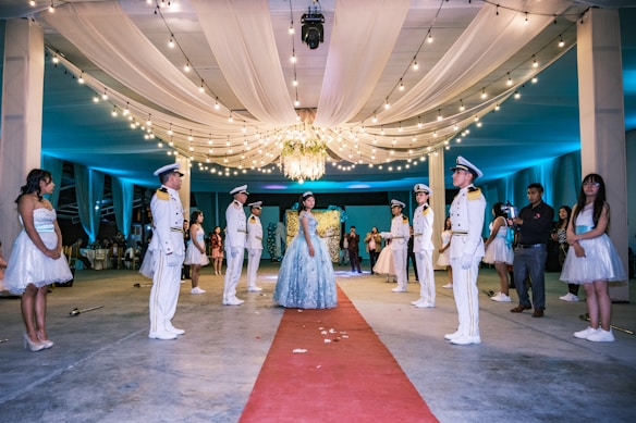 A formal event featuring a girl in an elegant blue gown standing on a red carpet, surrounded by uniformed individuals in white suits with hats. The setting is beautifully decorated with draped white fabric on the ceiling and an abundance of string lights contributing to a festive atmosphere. A chandelier hangs centrally, illuminating the scene. Several other people dressed in formal attire stand on either side, observing the moment.
