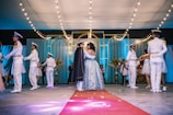 A formal dance event takes place in an elegantly decorated hall with string lights and draped curtains. Several couples are dancing, one in the center appears to be the focus, wearing a detailed gown and a cape. Surrounding them are others dressed in formal white and gold uniforms or formal dresses.