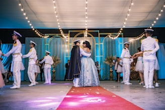 A formal dance event takes place in an elegantly decorated hall with string lights and draped curtains. Several couples are dancing, one in the center appears to be the focus, wearing a detailed gown and a cape. Surrounding them are others dressed in formal white and gold uniforms or formal dresses.