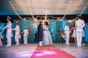 A formal dance event takes place in an elegantly decorated hall with string lights and draped curtains. Several couples are dancing, one in the center appears to be the focus, wearing a detailed gown and a cape. Surrounding them are others dressed in formal white and gold uniforms or formal dresses.