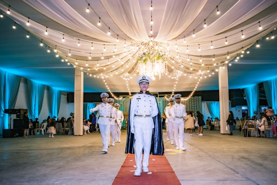 A group of people in naval uniforms are walking down a red carpet inside an elegantly decorated hall. The hall features draped white fabric on the ceilings and is lit with string lights, creating an ambient atmosphere. The background shows several seated guests and tables.