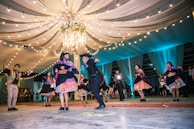A group of people dressed in vibrant, traditional attire are dancing energetically on a decorated floor. Above them, a canopy of draped fabric and string lights creates a festive atmosphere. The dancers appear to be in a celebratory mood, possibly participating in a cultural event or celebration.