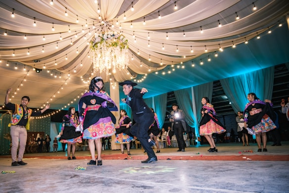 A group of people dressed in vibrant, traditional attire are dancing energetically on a decorated floor. Above them, a canopy of draped fabric and string lights creates a festive atmosphere. The dancers appear to be in a celebratory mood, possibly participating in a cultural event or celebration.