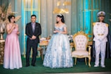 A formal event with four people standing on a decorated stage. A young woman in a blue floral gown and tiara stands next to a gold-trimmed chair. To her left, a man in a suit and tie is standing with his hands together. Next to him, a woman in a pink sparkly dress holds a microphone. On her right, a man in a white naval uniform with a gold shoulder trim and hat stands at attention. The background features decorative lighting and the words '15 años'.
