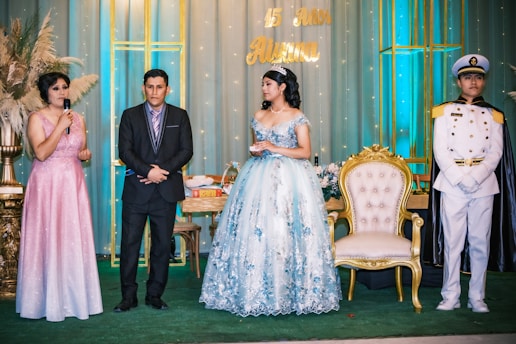 A formal event with four people standing on a decorated stage. A young woman in a blue floral gown and tiara stands next to a gold-trimmed chair. To her left, a man in a suit and tie is standing with his hands together. Next to him, a woman in a pink sparkly dress holds a microphone. On her right, a man in a white naval uniform with a gold shoulder trim and hat stands at attention. The background features decorative lighting and the words '15 años'.