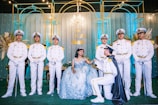 A young woman in a blue, intricately designed gown and tiara is seated surrounded by six uniformed naval officers. One of the officers is kneeling and holding her hand, with the others standing in a formal pose. The background features elegant decor with floral arrangements and soft blue lighting.