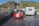 A red vintage sports car with white racing stripes is parked on a ferry along with other vehicles, including a camper van and a bicycle. The ferry is crossing a river, with a scenic view of hills and a castle in the background. The water is calm, and there are buildings along the riverbank.