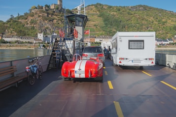 A red vintage sports car with white racing stripes is parked on a ferry along with other vehicles, including a camper van and a bicycle. The ferry is crossing a river, with a scenic view of hills and a castle in the background. The water is calm, and there are buildings along the riverbank.