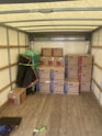 An empty truck interior with wooden floorboards is visible. Several cardboard boxes labeled 'Lowe's' are stacked neatly along the back of the truck. A green-wrapped item, possibly furniture, is secured at the side with a moving blanket partially covering it. There is a black and blue moving blanket and a green strap on the ground.