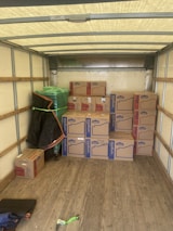 An empty truck interior with wooden floorboards is visible. Several cardboard boxes labeled 'Lowe's' are stacked neatly along the back of the truck. A green-wrapped item, possibly furniture, is secured at the side with a moving blanket partially covering it. There is a black and blue moving blanket and a green strap on the ground.