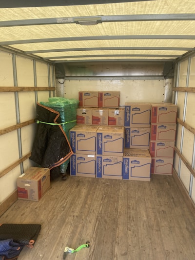 An empty truck interior with wooden floorboards is visible. Several cardboard boxes labeled 'Lowe's' are stacked neatly along the back of the truck. A green-wrapped item, possibly furniture, is secured at the side with a moving blanket partially covering it. There is a black and blue moving blanket and a green strap on the ground.
