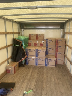 An empty truck interior with wooden floorboards is visible. Several cardboard boxes labeled 'Lowe's' are stacked neatly along the back of the truck. A green-wrapped item, possibly furniture, is secured at the side with a moving blanket partially covering it. There is a black and blue moving blanket and a green strap on the ground.