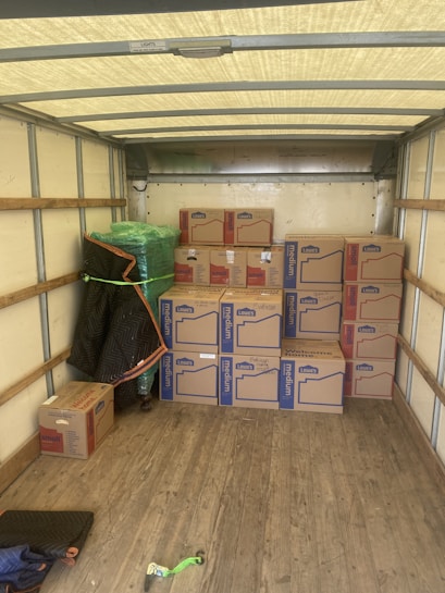 An empty truck interior with wooden floorboards is visible. Several cardboard boxes labeled 'Lowe's' are stacked neatly along the back of the truck. A green-wrapped item, possibly furniture, is secured at the side with a moving blanket partially covering it. There is a black and blue moving blanket and a green strap on the ground.