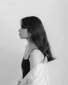 A monochrome profile portrait of a young woman with long, wavy hair. She is wearing a sleeveless top and an open shirt draped over her shoulders. The background is plain and light, emphasizing the subject's silhouette.