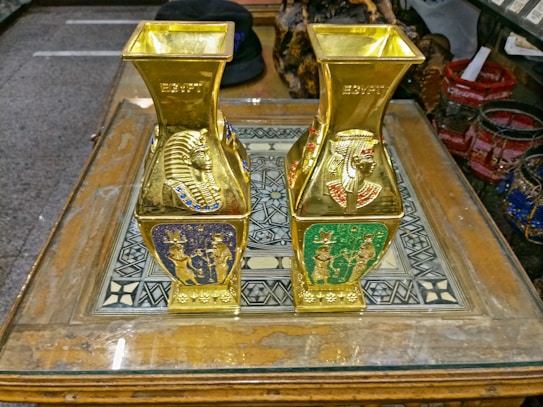Two ornate golden vases with Egyptian motifs, including pharaoh heads and hieroglyphic designs, are placed on an intricate wooden table with geometric patterns. The vases have vibrant red, blue, and green ornamental detailing.