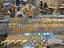 A vibrant market stall filled with an array of intricately designed decorative items. The shelves display various sculptures, including animals like elephants and tigers, as well as human figures and abstract art. The objects are detailed with gold, blue, and colorful patterns, creating a visually rich and diverse collection.
