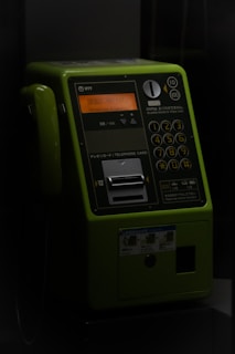 A green public telephone booth with a numeric keypad and a slot for a telephone card. The lighting is dim, giving the telephone a dark and slightly retro appearance. The phone has buttons with numbers, symbols and instructions written in Japanese. An orange display screen can be seen in the middle part, above the keypad. The handset is placed on the left side of the phone.