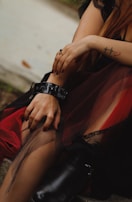 A person is sitting with their hands resting on their legs. They have a tattoo of a heartbeat with a cross and heart on their arm. They are wearing a black dress with red and mesh layers, complemented by black boots with high heels. A black ring is on one finger, and the nails are painted dark. The setting appears outdoors, with a stone or concrete surface nearby.