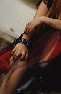 A person is sitting with their hands resting on their legs. They have a tattoo of a heartbeat with a cross and heart on their arm. They are wearing a black dress with red and mesh layers, complemented by black boots with high heels. A black ring is on one finger, and the nails are painted dark. The setting appears outdoors, with a stone or concrete surface nearby.