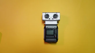 A portable field audio recorder with a small display, buttons, and two large knobs is placed against a bright yellow background.