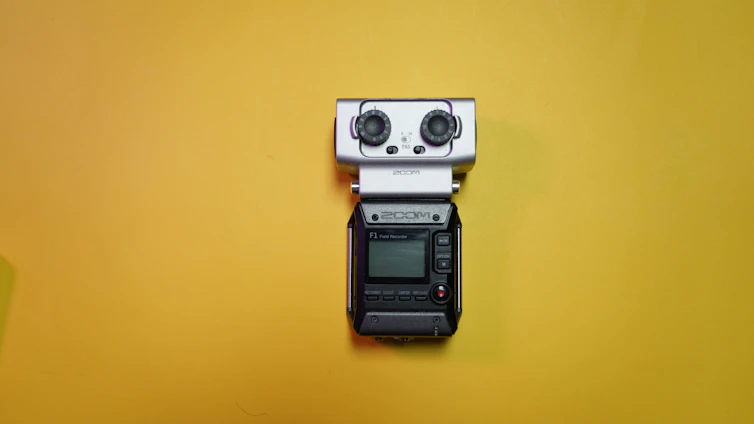 A portable field audio recorder with a small display, buttons, and two large knobs is placed against a bright yellow background.