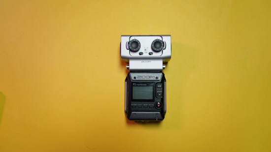 A portable field audio recorder with a small display, buttons, and two large knobs is placed against a bright yellow background.