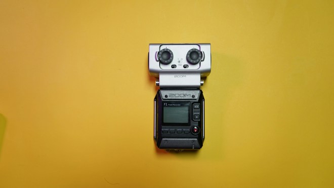A portable field audio recorder with a small display, buttons, and two large knobs is placed against a bright yellow background.