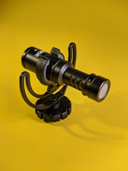 A black microphone with a sturdy mount against a vibrant yellow background. The microphone features brand labeling and is positioned upright, casting a faint shadow.