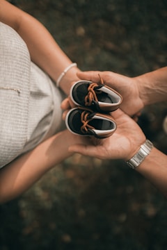 A person holding a pair of small baby shoes with tan laces in their hands. The hands are positioned over a blurred background featuring grass and some scattered leaves. The person holding the shoes is wearing a light-colored dress, and another hand is visible, wearing a watch.