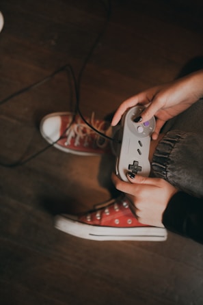 Hands holding a retro game controller, with visible red sneakers and a wooden floor in the background.